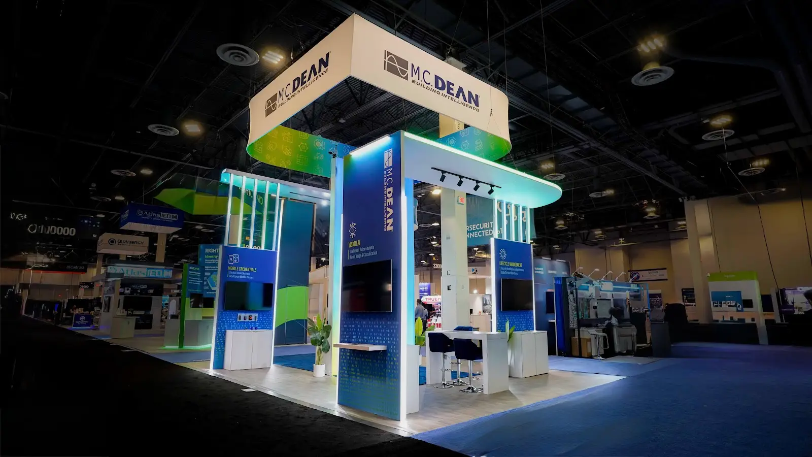 M.C. Dean 20x30 island exhibit at ISC West 2026 — front-left view showing all four navy kiosk towers, backlit overhead canopy, and cyan LED tube columns at entry