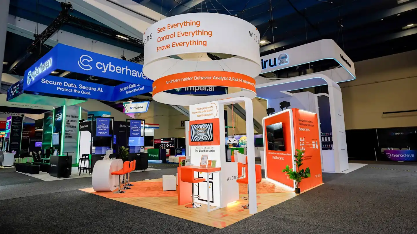 Front-right diagonal perspective of the WE3DS 20x20 ft island booth at RSA Conference 2026 showing the right exterior vivid orange informational panel with bold white typography reading Beyond endpoint limitations Visibility at your systems core and the BlackBox Series flow diagram illustrating the path from User icons through Application Server to DB Server demonstrating the platform detection architecture, the double-ring cylindrical hanging sign visible overhead with the outer white ring and inner orange band both internally backlit, white pod egg-style modern lounge chairs in the foreground providing comfortable seating for extended conversations, the blond oak raised platform flooring creating a warm residential contrast against the dark convention floor, monitors displaying the signature concentric spiral vortex pattern, orange bar stools at high-top tables, cyan LED accent strips along the white structural arch frames, and the neighboring Cyberhaven booth with its distinctive blue branding visible in the background to the right confirming the South Expo Hall positioning at RSA Conference 2026 Moscone Center