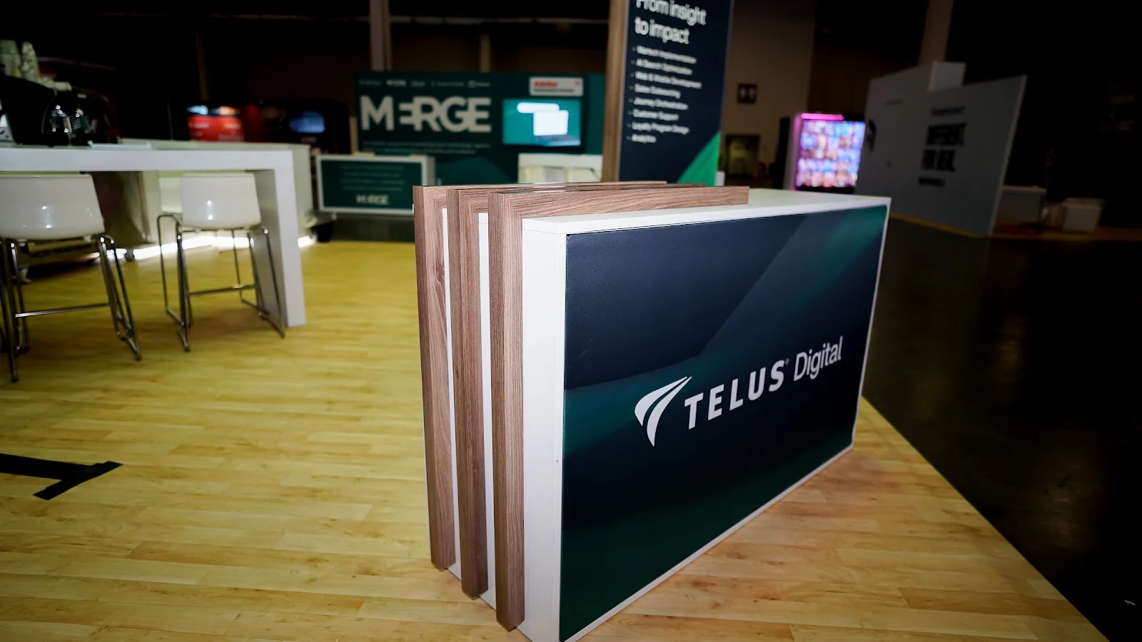 Close-up of TELUS Digital reception counter showing layered alternating walnut wood and white laminate architectural spine detail, dark navy branded front face with TELUS Digital logo, and white top surface at Adobe Summit 2026