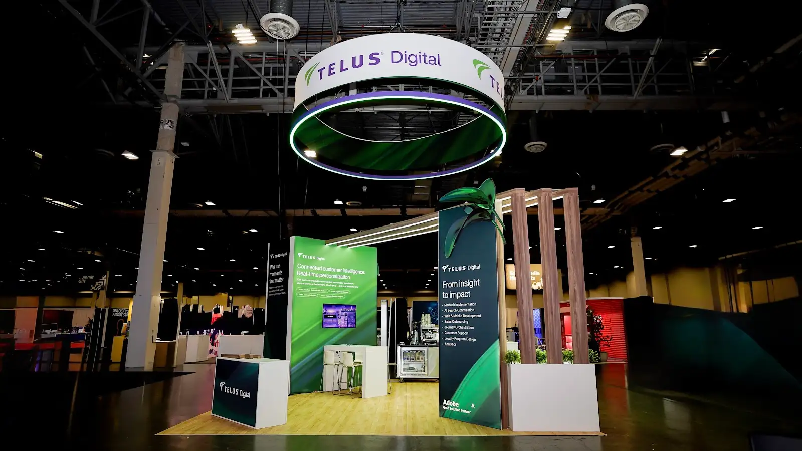 TELUS Digital 20x20 island exhibit at Adobe Summit 2026 — full front view showing circular LED halo hanging sign, vivid green Connected customer intelligence panel, and dark Build loyalty panel with vertical LED stripes at The Venetian Las Vegas