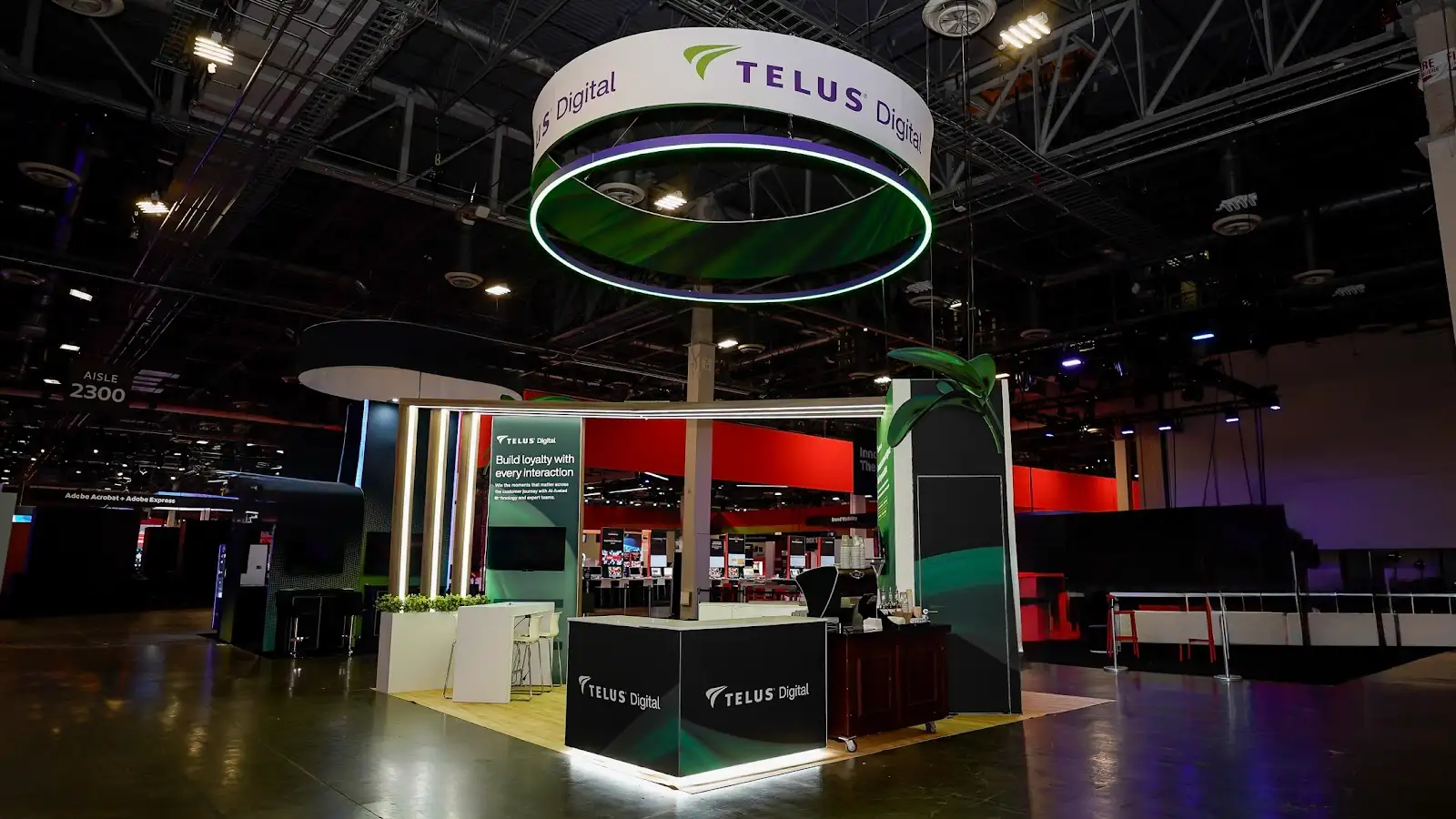 TELUS Digital booth Build loyalty face showing circular LED halo hanging sign glowing in dark convention hall, dark charcoal panel with vertical mint and teal LED stripes, and white planters with green topiary at Adobe Summit 2026