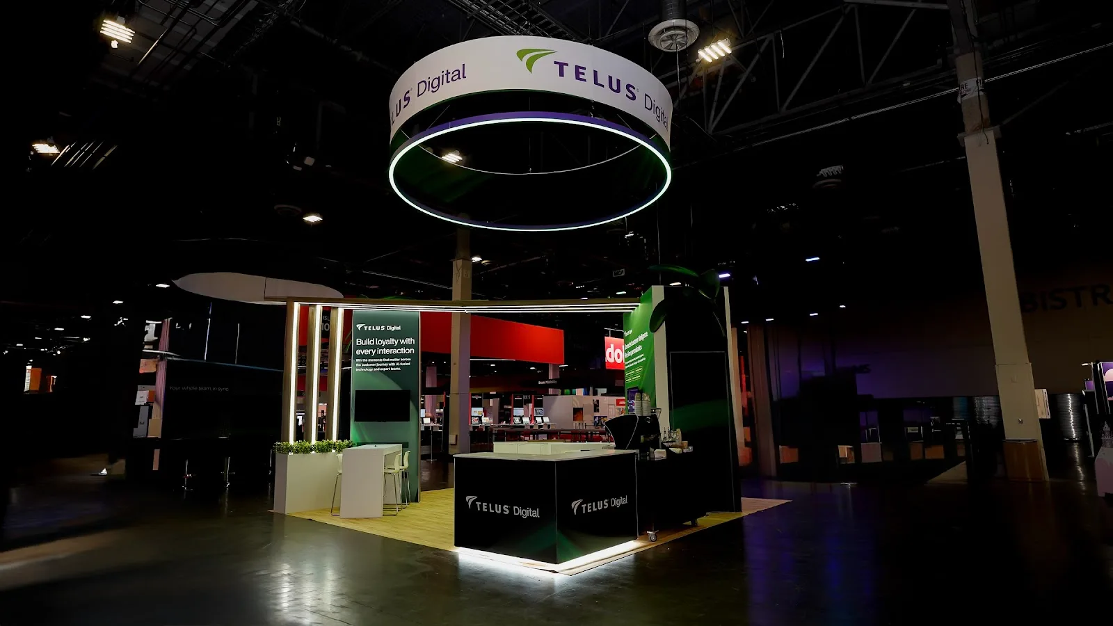TELUS Digital booth environmental shot in dark convention hall showing lighthouse effect of circular LED ring against ceiling rigging, dark panel with vertical LED stripes, and maple platform glowing warm against dark concrete floor at Adobe Summit 2026
