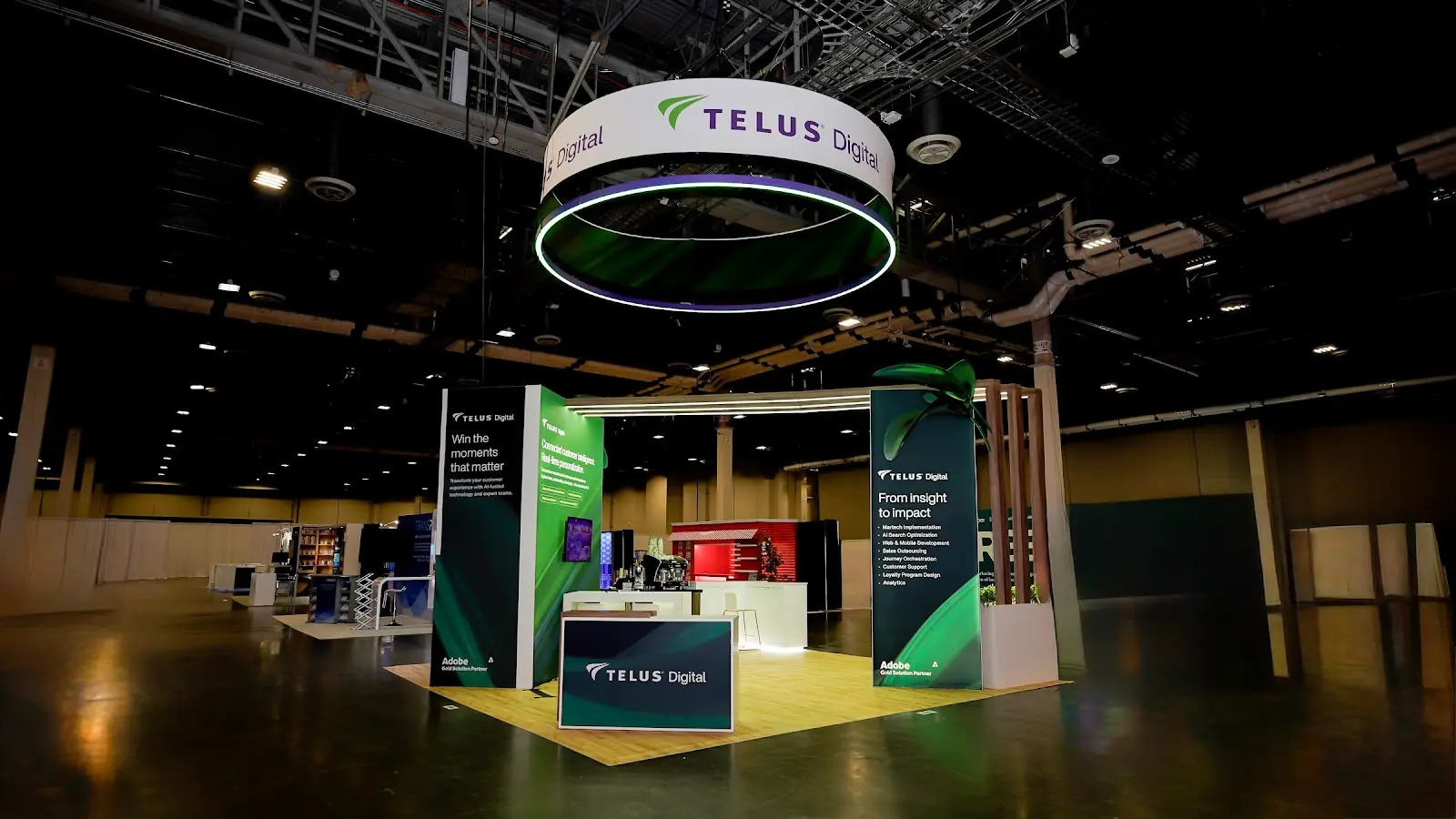 TELUS Digital booth alternate deployment showing front counter with outward-facing kiosk monitor, green panel, dark panel, hanging sign, and red interior hospitality bar accent in ochre-walled convention venue