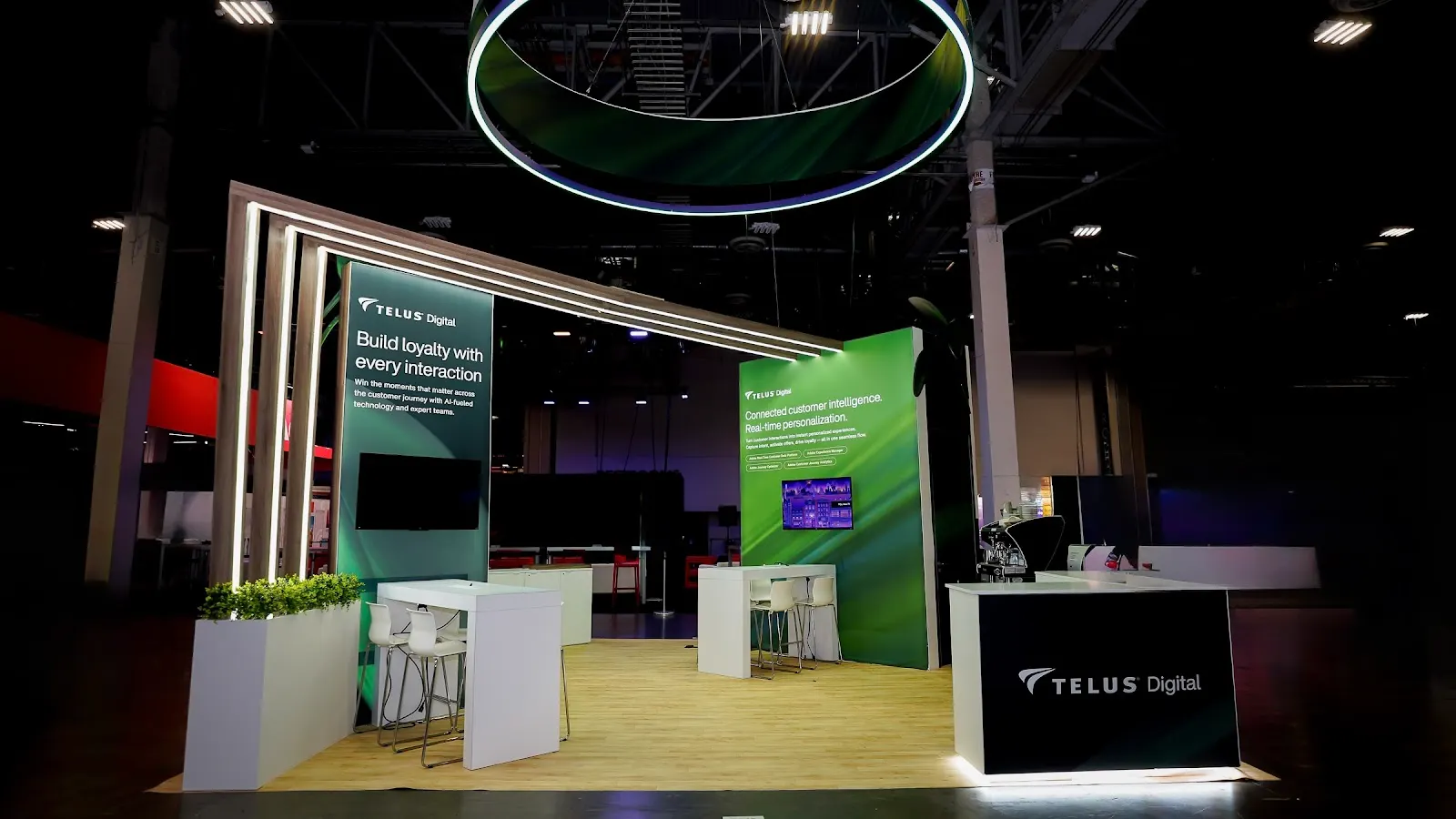 TELUS Digital booth interior shot showing LED portal gateway frame with horizontal LED bars, Build loyalty dark panel with vertical LED stripes on left, Connected customer intelligence green panel with live UI demo on right, and white planters with green topiary at Adobe Summit 2026