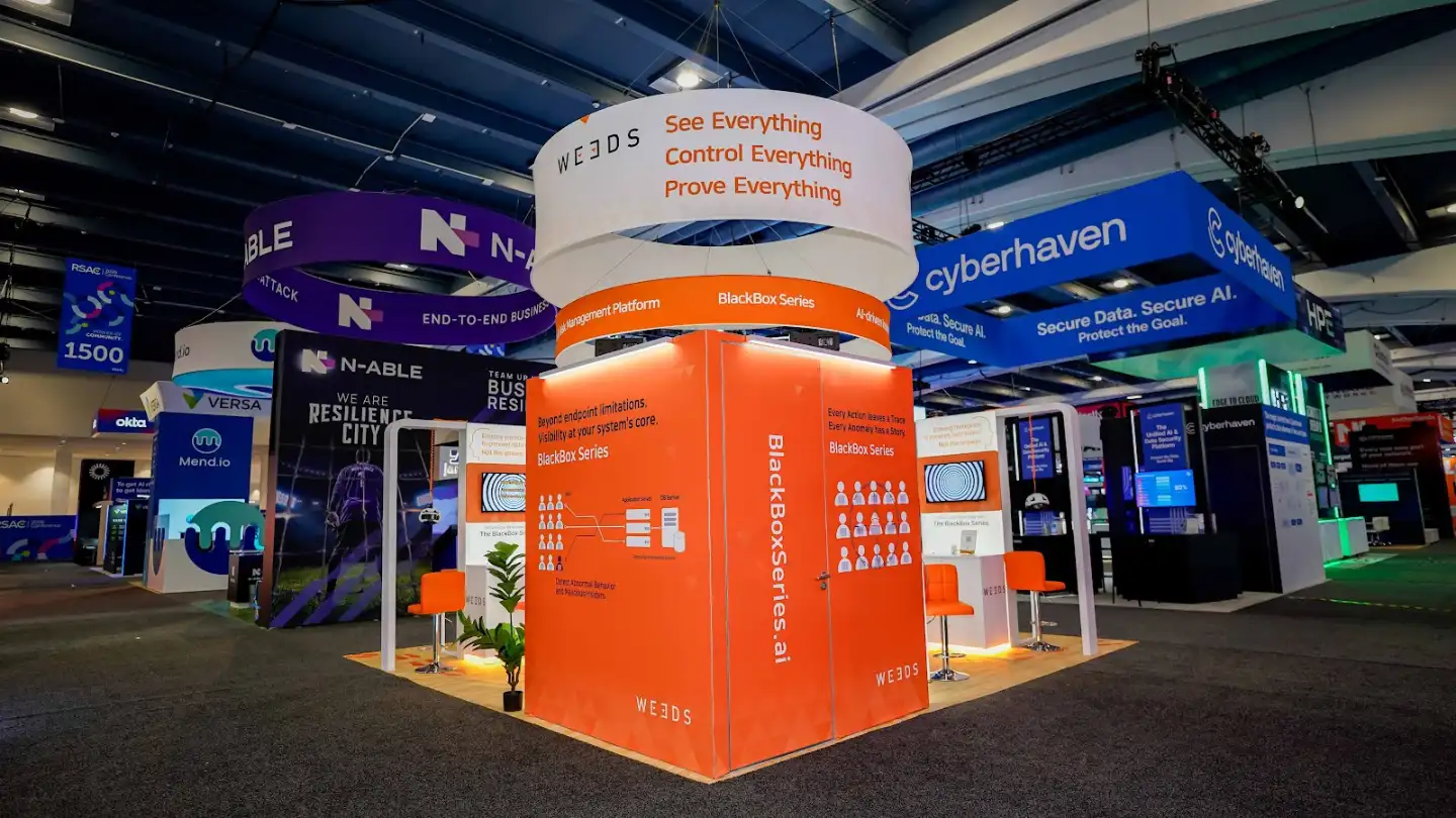 Rear view of the WE3DS 20x20 ft island booth at RSA Conference 2026 showing the symmetrical dual-panel back wall with the left panel reading Beyond endpoint limitations Visibility at your systems core with embedded 55-inch monitor displaying the concentric spiral animation and the right panel reading Every Action leaves a Trace Every Anomaly has a Story with its own embedded monitor and user-icon workforce visualization graphic, the tall white central pylon between both panels displaying BlackBoxSeries.ai in vertical full-height typography, the double-ring cylindrical hanging sign visible overhead from behind, RSAC Conference signage and event branding confirming the venue, neighboring Cyberhaven booth with blue branding visible to the left, N-able booth with purple branding visible to the right, HPE and Okta booth signage in the background, blond oak raised platform, white structural frames bordering each orange panel, the entire rear composition creating a symmetrical triptych of product identity and messaging visible to attendees approaching from the rear aisle corridor