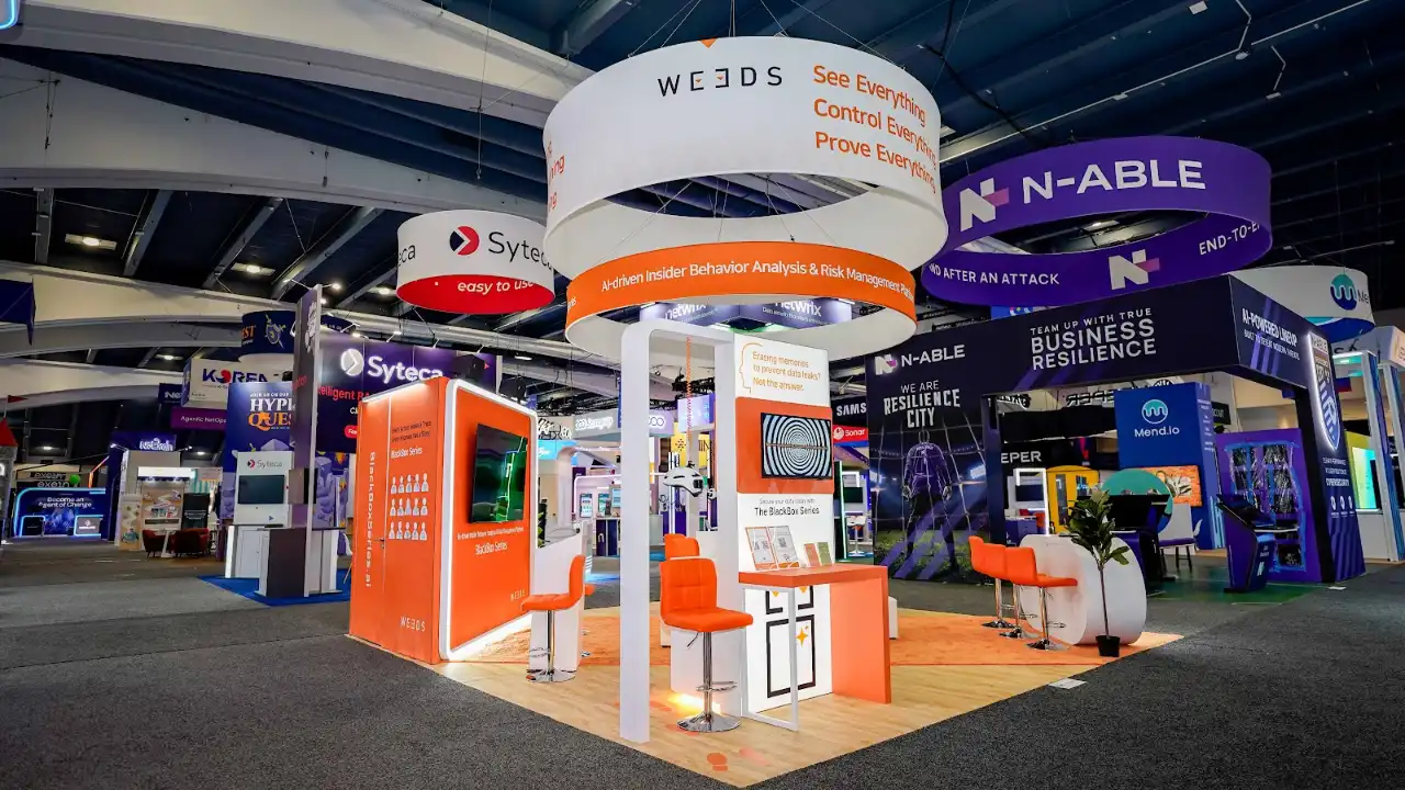 Front-center comprehensive wide-angle view of the WE3DS 20x20 ft island exhibit at RSA Conference 2026 Moscone Center South Expo Hall showing the full booth from the primary aisle approach with the left exterior vivid orange panel displaying the Erasing memories to prevent data leaks Not the answer problem statement and QR code, the central QR kiosk visible inside the booth footprint, the right exterior panel partially visible with the Beyond endpoint limitations messaging, the double-ring cylindrical hanging sign overhead with the outer white ring and inner orange band both internally backlit, the blond oak raised wood-grain platform flooring spanning the entire island, cyan teal LED accent strips along the white portal arch frames, the white oval reception desk with WE3DS branding at center-front, orange bar stools and white high-top tables in the interior, monitors displaying the signature black-and-white concentric spiral vortex pattern, fiddle-leaf fig potted plants adding organic warmth, an orange floor dot graphic at the platform edge, and the neighboring Syteca N-able and Mend.io booths visible in the background confirming the South Expo Hall RSA Conference 2026 environment with dark convention ceiling and overhead rigging