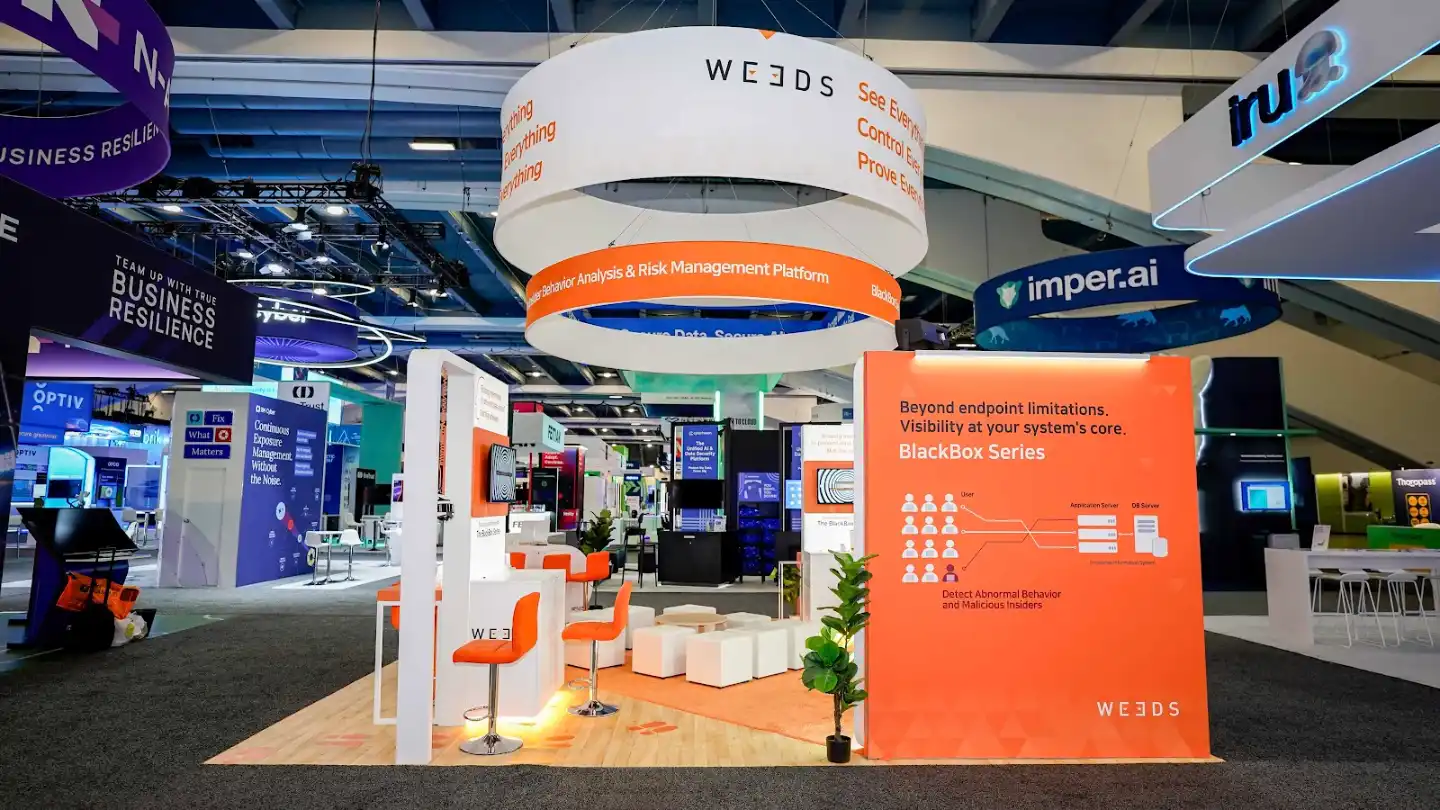 Rear-left angle of the WE3DS 20x20 island booth showing the right exterior vivid orange panel with detailed infographic messaging in white typography, the double-ring overhead sign from behind, blond oak platform flooring, white structural frames bordering the panel, neighboring N-able purple booth and Dr. Cyber booth visible in background, RSA Conference Moscone Center South Expo Hall environment