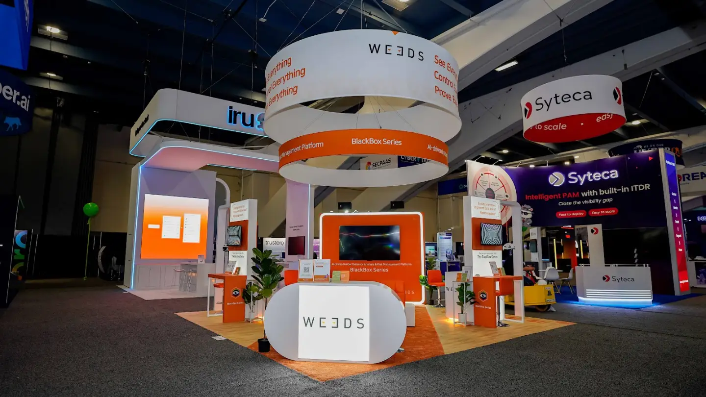 Wide front-left 45-degree angle establishing shot of the complete WE3DS 20x20 ft island exhibit at RSA Conference 2026 in the Moscone Center South Expo Hall San Francisco showing the dramatic double-ring cylindrical internally-lit hanging sign suspended from the convention ceiling with the outer white ring displaying the WE3DS logo and orange chevron on one face and the bold orange See Everything Control Everything Prove Everything manifesto on the opposite face and the inner smaller orange band ring with white text reading Ai-driven Insider Behavior Analysis and Risk Management Platform BlackBox Series, the white oval pill-shaped reception desk centered at the front of the booth with WE3DS branding on its face, vivid orange exterior informational panels with white typography on all visible sides, blond oak wood-grain raised platform flooring throughout, cyan teal LED accent strips running along the white portal arch frame edges adding a tech-forward contrast to the orange palette, three monitors displaying the signature black-and-white concentric spiral vortex pattern that is the BlackBox Series visual identity, orange cushioned chrome-base bar stools, fiddle-leaf fig potted plants in black planters adding organic warmth, white high-top tables, and the neighboring Syteca booth visible in the background confirming the South Expo Hall RSA Conference 2026 environment