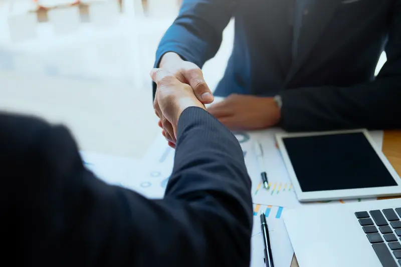 Business professionals shaking hands during a meeting to finalize a trade show exhibit booth partnership, with charts, a tablet, and a laptop on the table. Sponsorship vs. Exhibiting Image.webp
