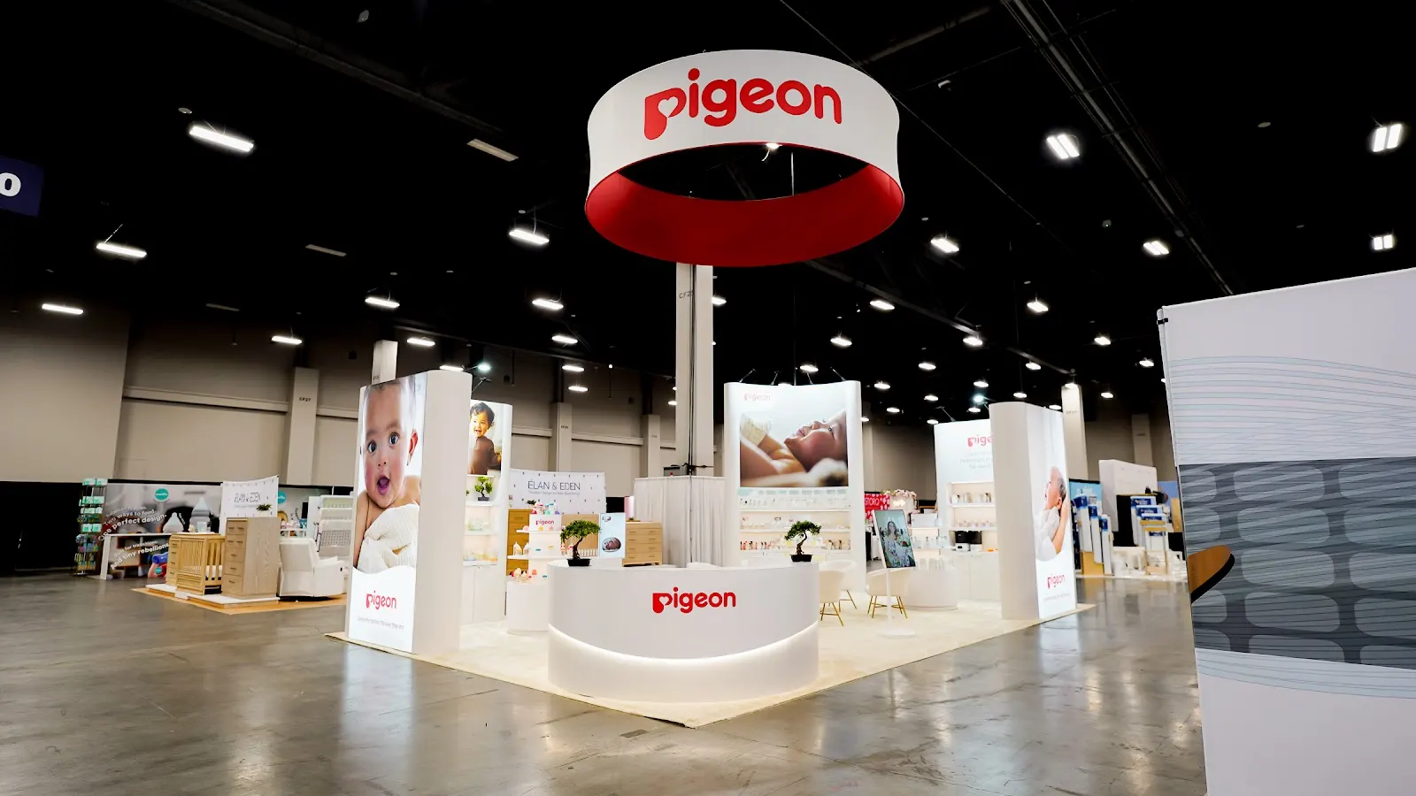 Pigeon baby products 20x30 custom island booth at ABC Kids Expo 2026 in Mandalay Bay Convention Center Las Vegas, featuring overhead hanging brand sign, open layout with white product shelving along perimeter walls, interactive demo stations with warm directional lighting, and raised platform highlighting flagship feeding and skincare collections