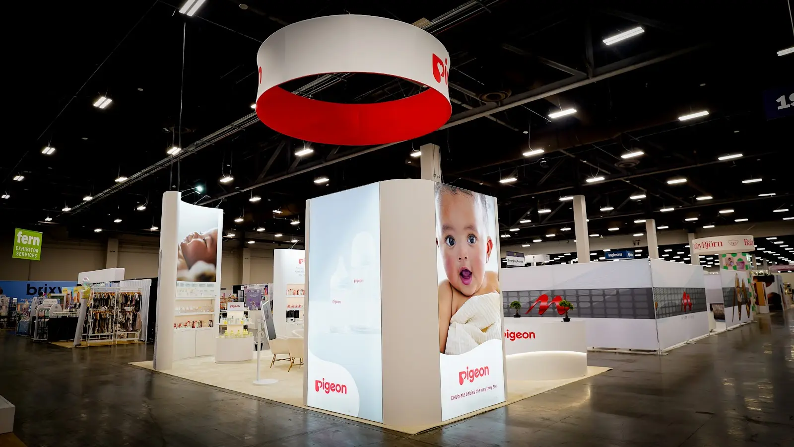 Full-length perspective view of the Pigeon baby products custom island booth at ABC Kids Expo 2026 at Mandalay Bay Las Vegas, showing the complete booth in active show conditions with visitors engaging with product stations, overhead hanging sign visible at height, warm interior lighting illuminating the white and blue brand environment, and the organized product display shelving running the full perimeter of the 600 square foot exhibit space