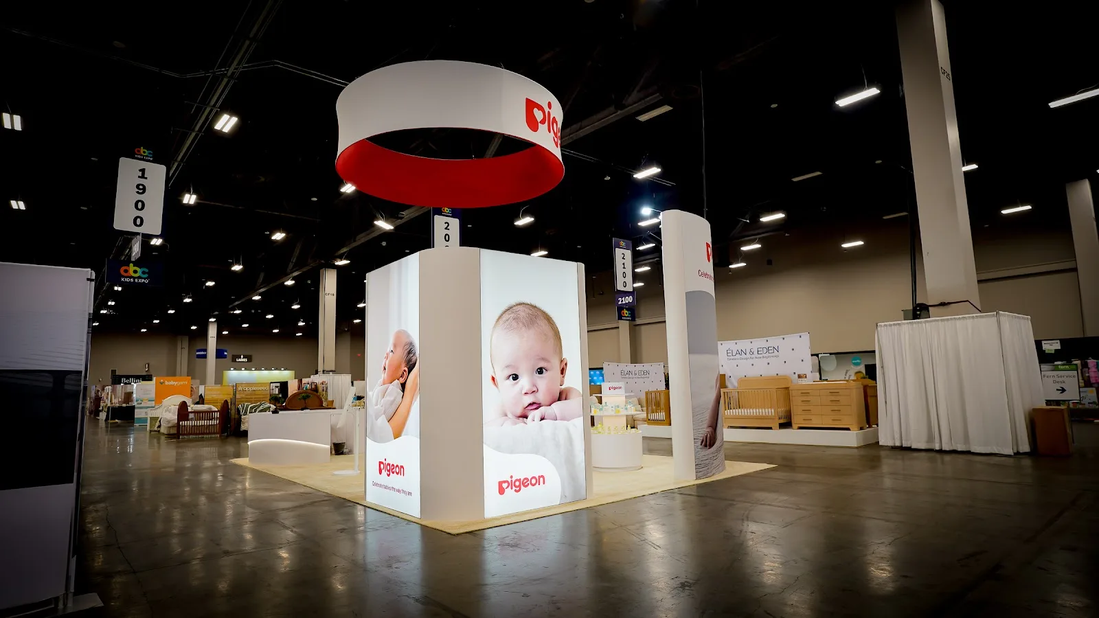 Exterior approach view of the Pigeon island exhibit at ABC Kids Expo 2026 showing the open perimeter layout with wide aisle access, prominent overhead hanging sign in Pigeon blue and white visible from across the convention hall, and the elevated raised platform section that defines the rear of the 20x30 booth structure