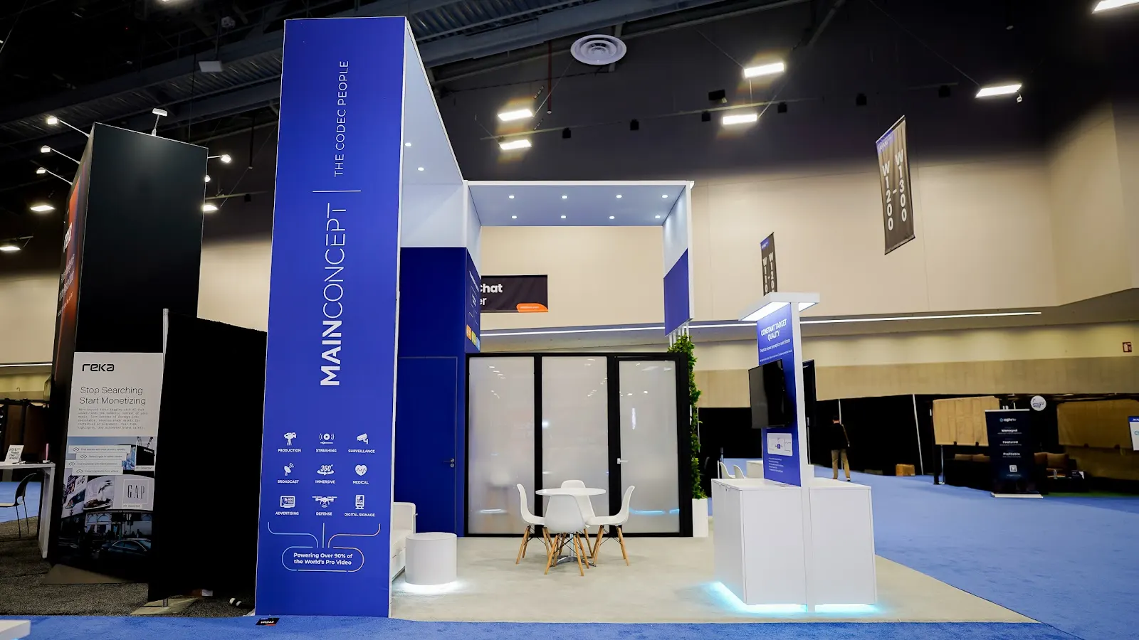 Full perspective view of the MainConcept blue and white custom island booth at NAB Show 2026 in the Las Vegas Convention Center, showing the complete technical zone layout with demonstration areas and open sightlines under active show conditions