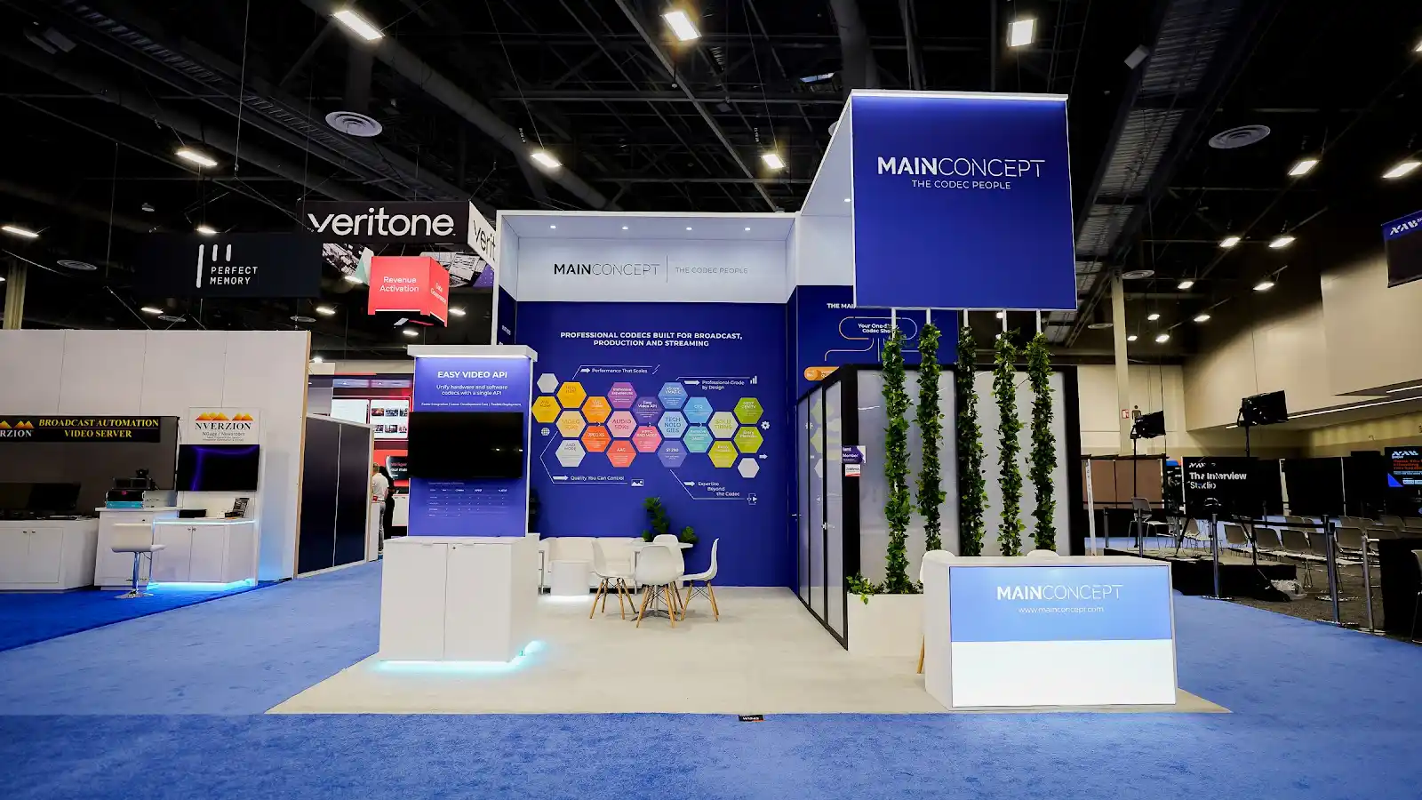 MainConcept 20x20 custom island booth at NAB Show 2026 in the Las Vegas Convention Center, front view showing the clean blue and white technical zone with open sightlines and precise geometric structure
