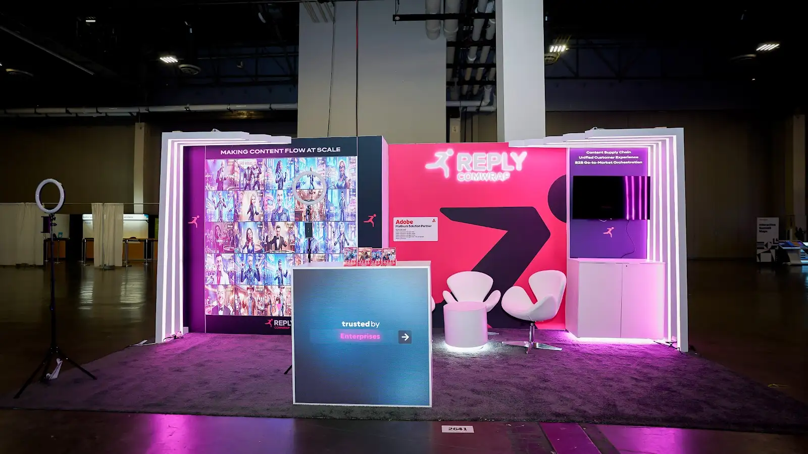Comwrap Reply booth at Adobe Summit 2026 seen from the aisle, with pink and purple illuminated panels framing the open entry into the exhibit