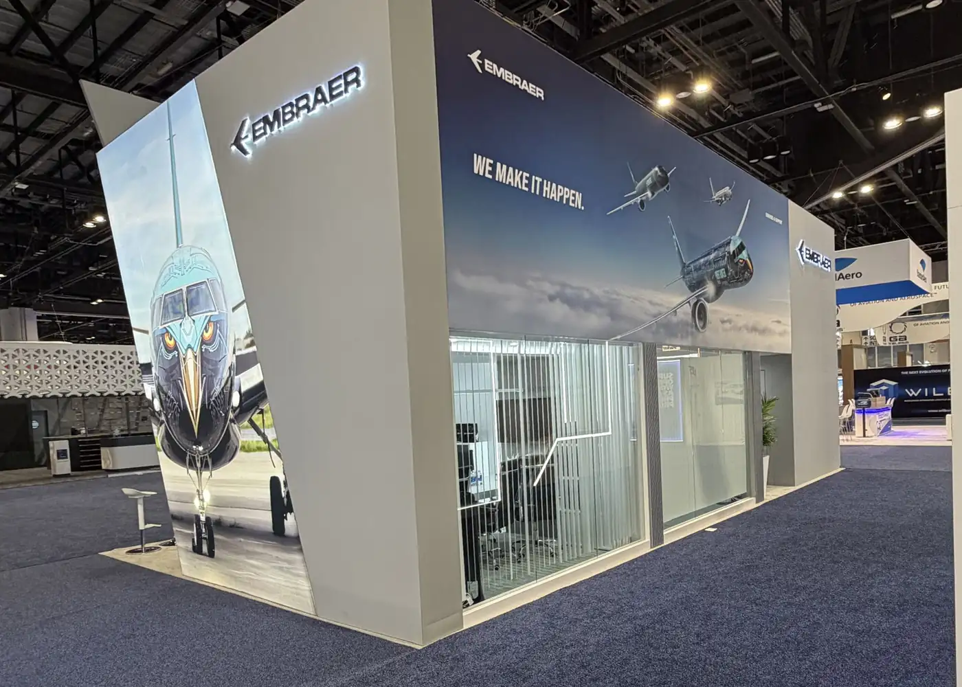 Corner of the Embraer double-deck booth at MRO Americas 2026 with the eagle-nose E-Jet livery graphic and glass meeting rooms beneath the flight mural