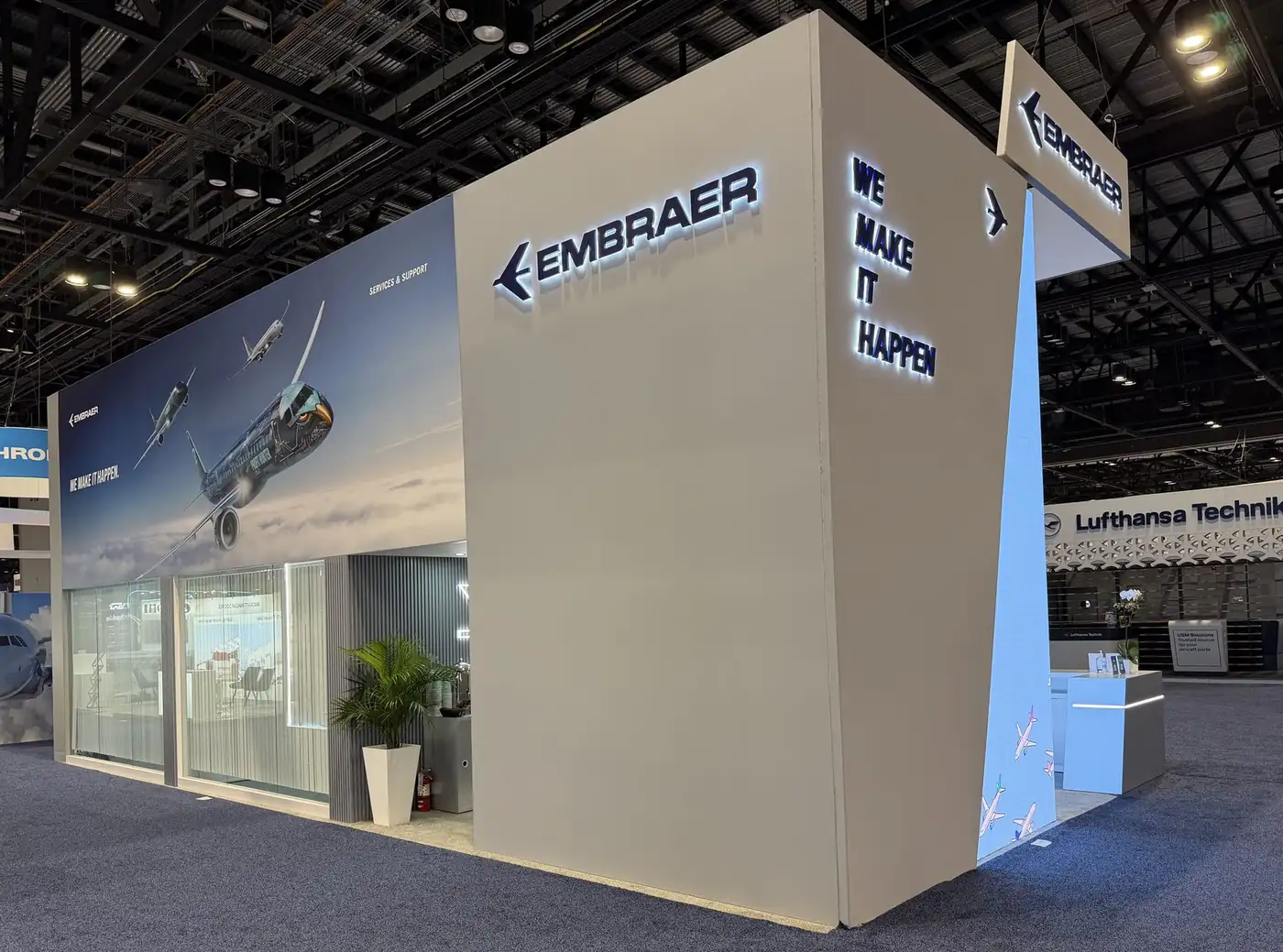 End wall of the Embraer booth at MRO Americas 2026 with backlit We Make It Happen dimensional letters and hanging sign