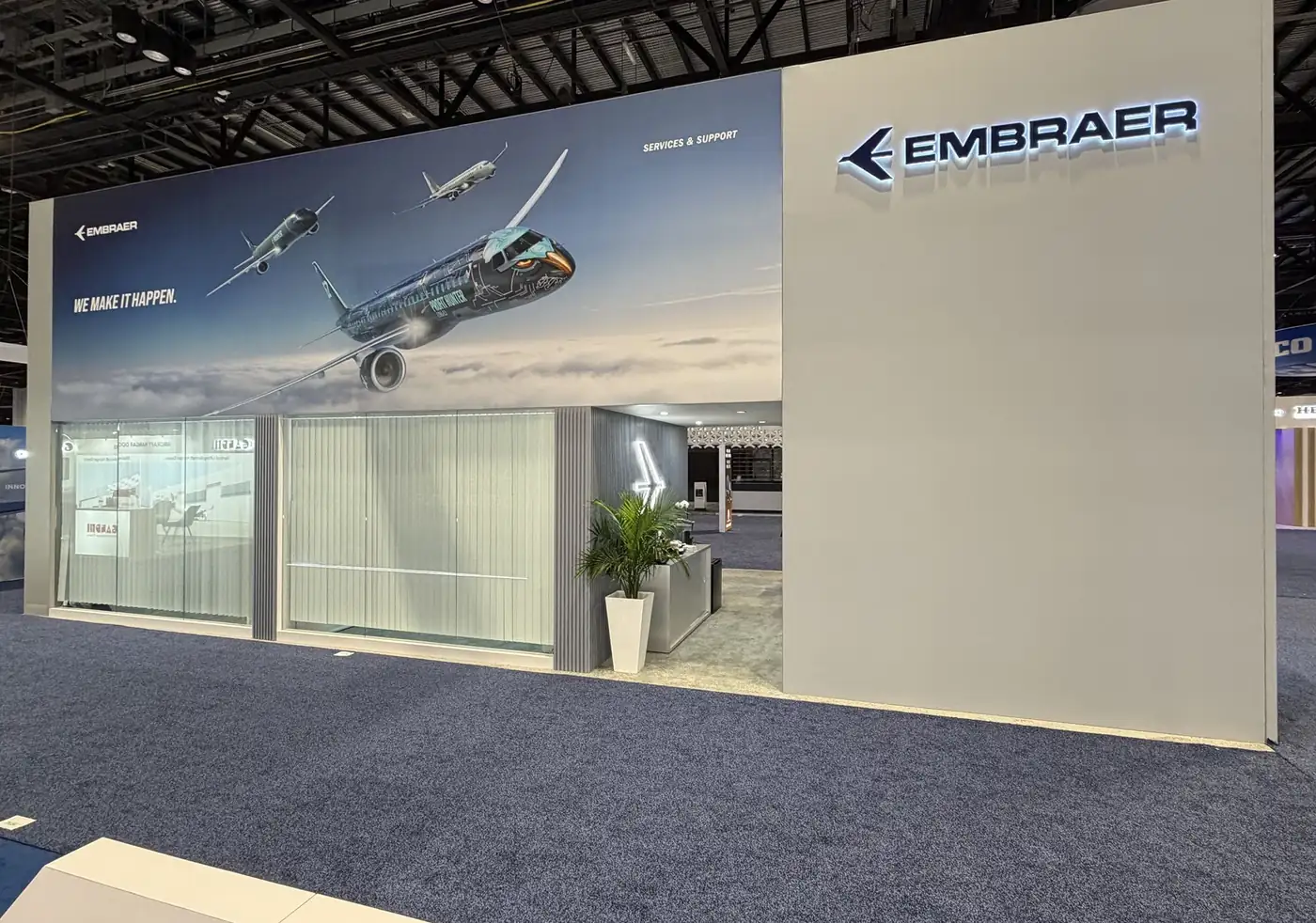 Side elevation of the Embraer double-deck booth at MRO Americas 2026 with E-Jet flight mural above ground-floor glass-walled meeting rooms