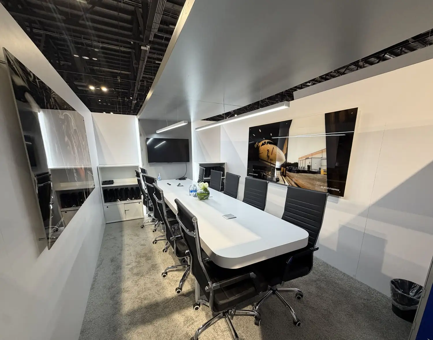 Upper-deck conference room inside the Embraer double-deck booth at MRO Americas 2026 with long white table, black leather chairs, and aviation photography