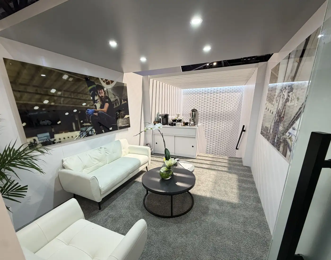 Upper-deck lounge in the Embraer double-deck booth at MRO Americas 2026 with white sofa, coffee bar, and Services and Support photography