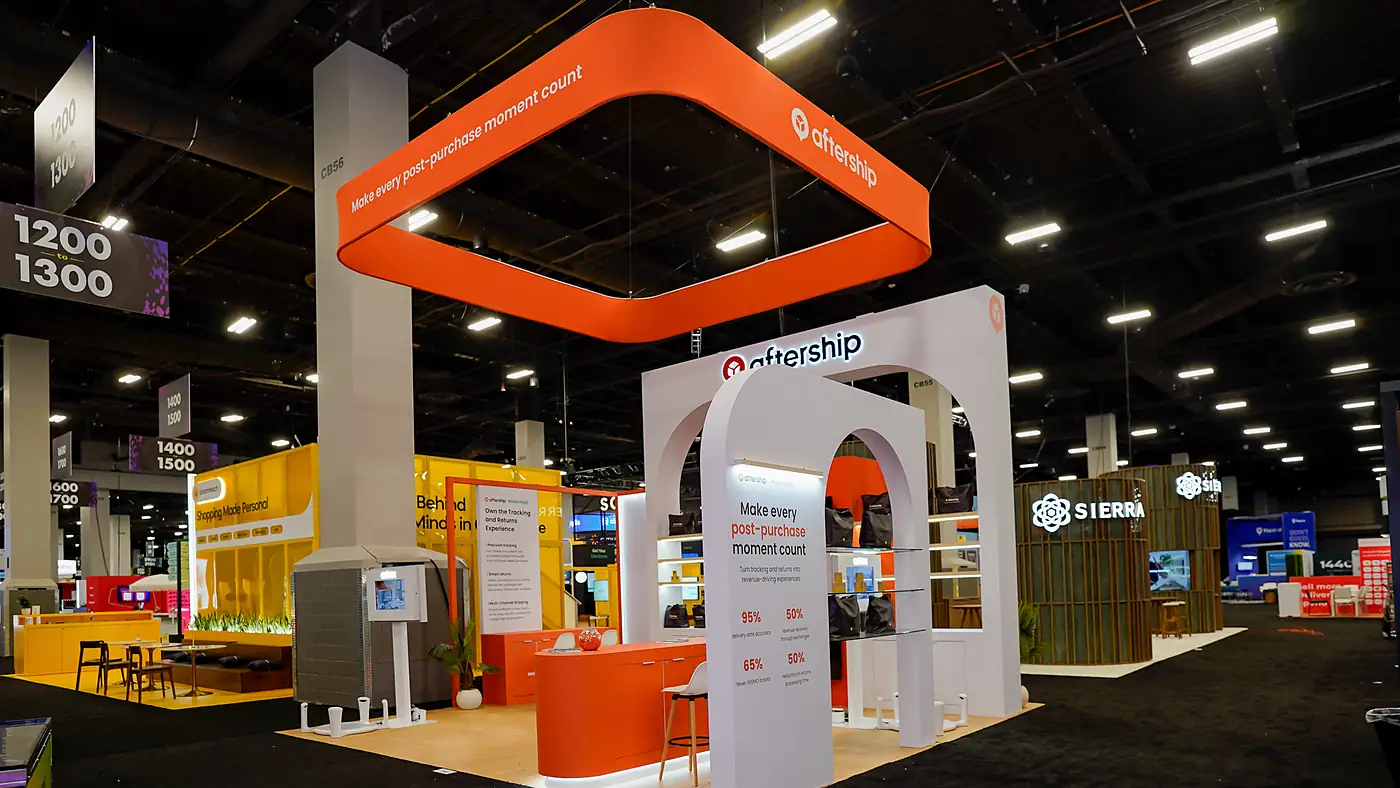 Aisle view of the AfterShip 20x20 island at Shoptalk Spring 2026 with the rounded-square orange hanging sign above white arched portals, an orange reception counter and a bar stool on light wood flooring