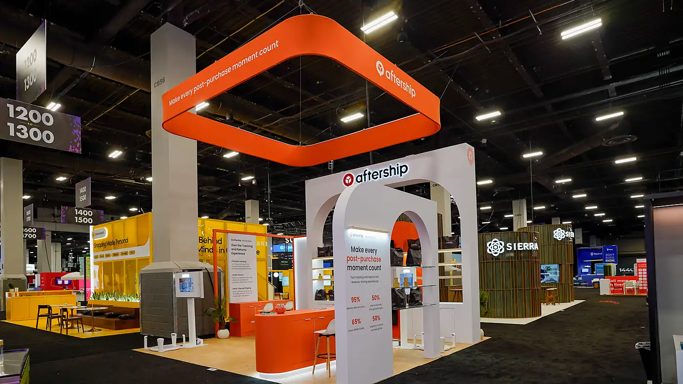 AfterShip 20x20 island booth at Shoptalk Spring 2026 with a rounded orange hanging sign reading Make every post-purchase moment count, white arched portals, an orange reception counter and backlit product shelves