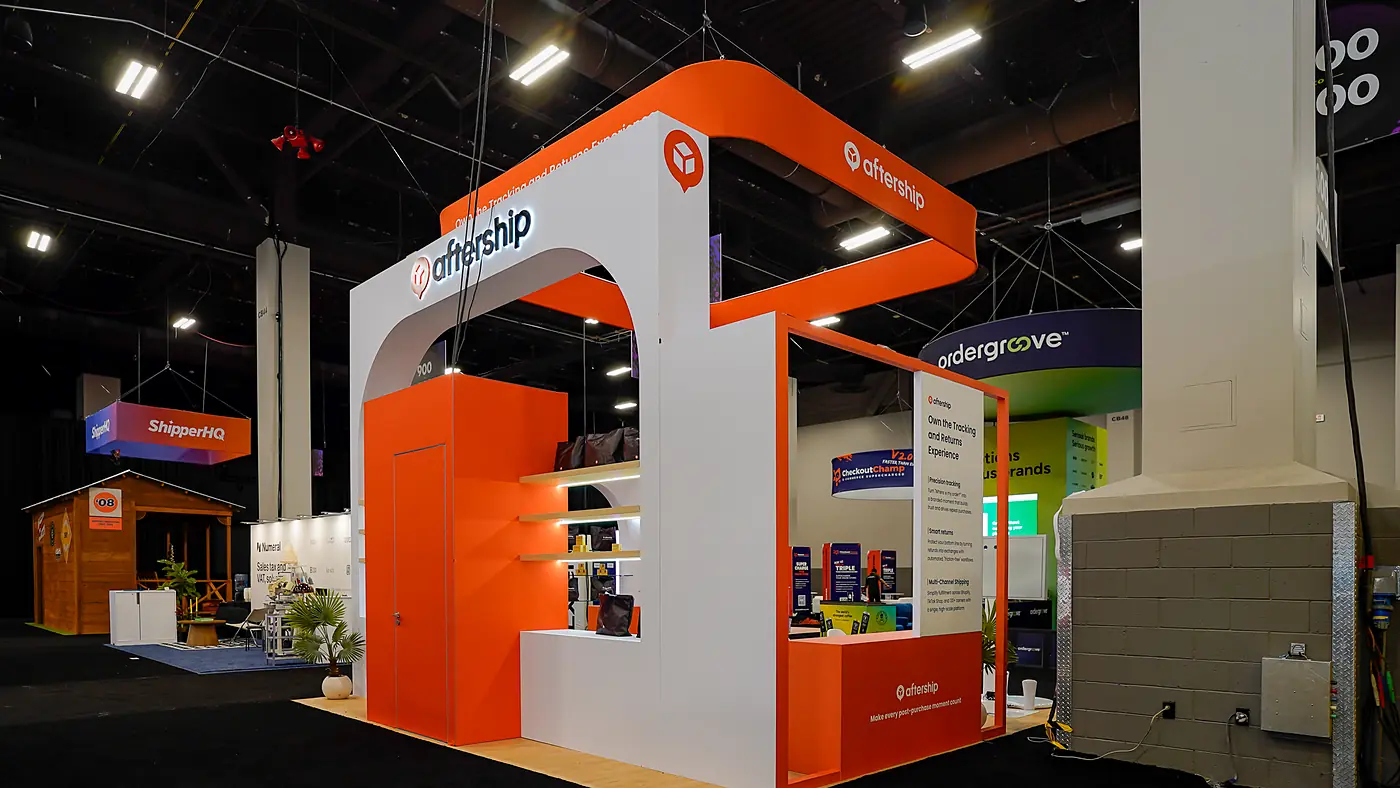 Rear corner of the AfterShip booth at Shoptalk Spring 2026 showing the orange hanging sign curving over a white arch, the illuminated AfterShip logo and the orange storage room beside a small arched messaging panel