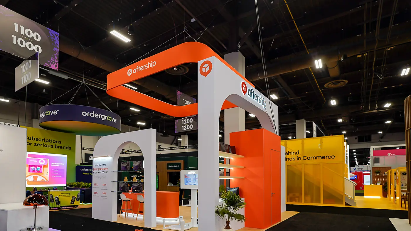 Side view of the AfterShip booth at Shoptalk Spring 2026 with backlit wood shelves of branded tote bags and product boxes, the orange storage room, the arch panel headline and the reception counter under the hanging sign