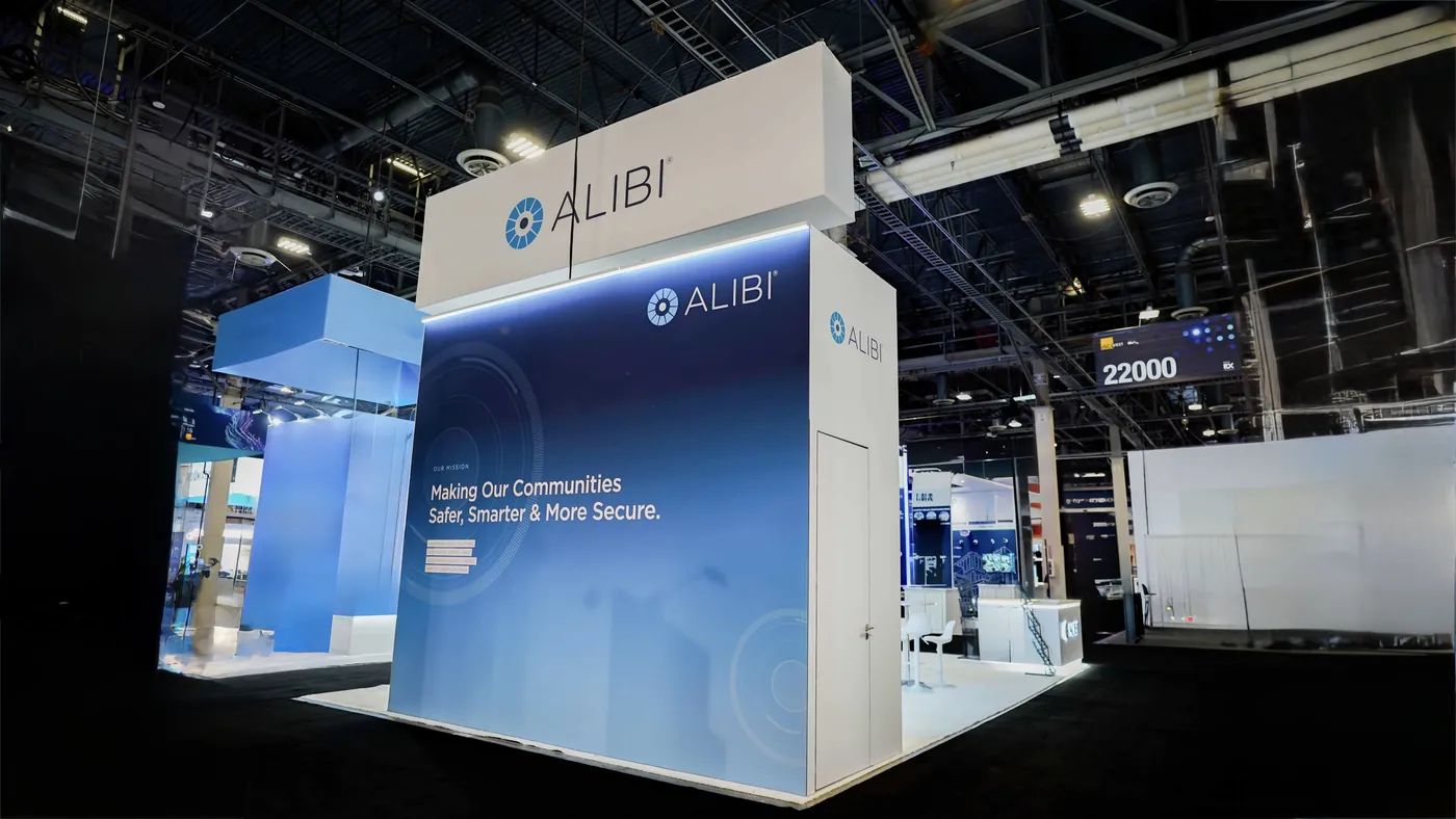 Side elevation of the Alibi Security booth at ISC West 2026 with the blue Our Mission graphic wall reading Making Our Communities Safer, Smarter and More Secure, a flush storage door and the ALIBI hanging sign above