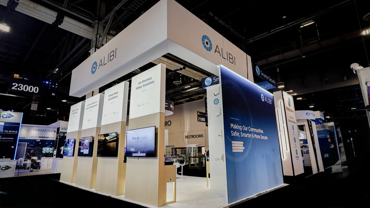 Aisle-side elevation of the Alibi Security 20x30 booth at ISC West 2026 showing four illuminated lightbox panels with monitors beneath, the white perimeter header with ALIBI wordmark and the blue mission wall