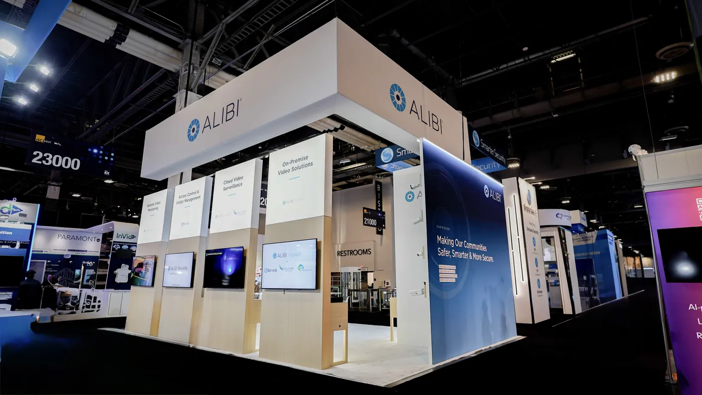 Rear corner view of the Alibi Security 20x30 island booth at ISC West 2026 showing the full white perimeter header, the four aisle-facing lightbox panels with screens and the blue mission wall