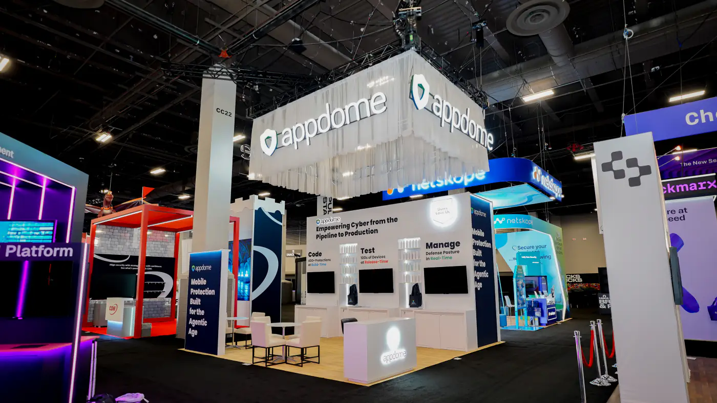 Appdome 20x20 island booth at Black Hat USA 2026 with a white sheer-draped hanging sign, Empowering Cyber from the Pipeline to Production back wall, three screens and white lounge seating