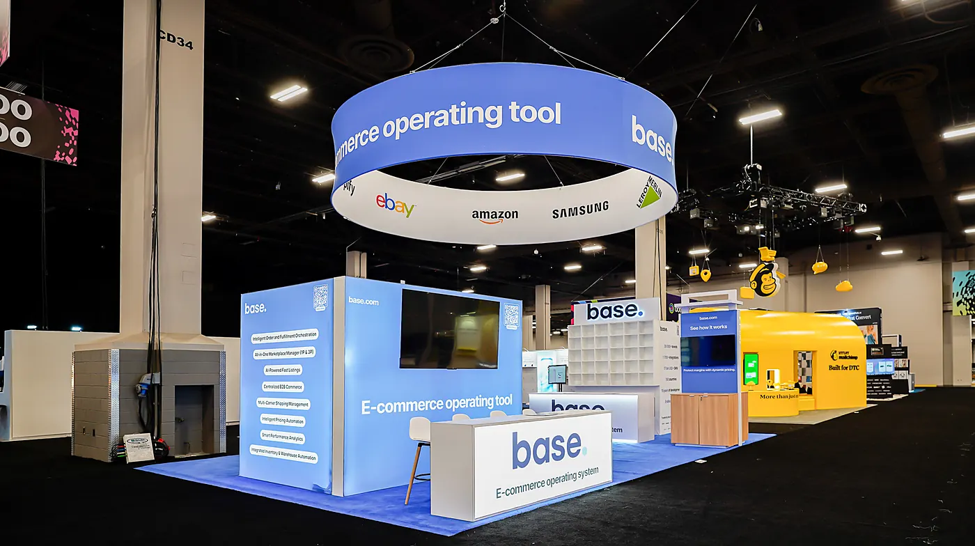 Base 20x30 booth at Shoptalk Spring 2026 showing the blue backlit graphic wall with feature list and monitor, white reception counter with Base logo, and the hanging ring sign with eBay, Amazon and Samsung logos
