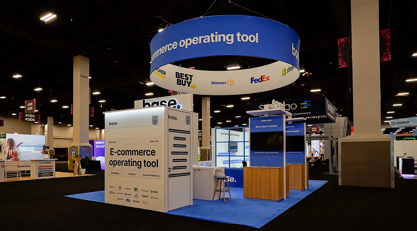 Base 20x30 island trade show booth at Shoptalk Spring 2026 seen from the aisle corner, with a blue circular hanging sign reading E-commerce operating tool and a backlit white feature tower listing Dropshipping, Wholesale and Resell partners