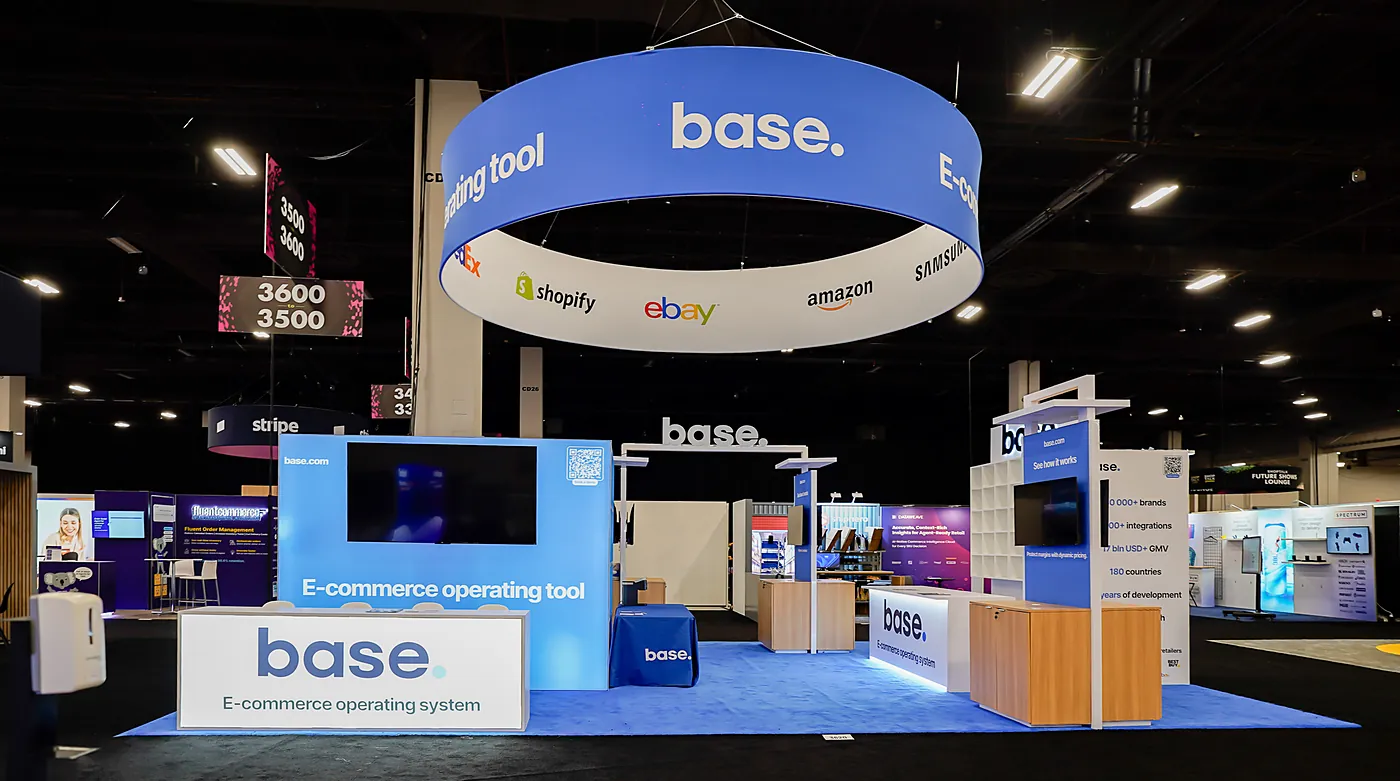Base 20x30 island booth at Shoptalk Spring 2026 from the open side, with the branded reception counter, blue graphic wall, See how it works kiosk and the large blue hanging ring sign overhead