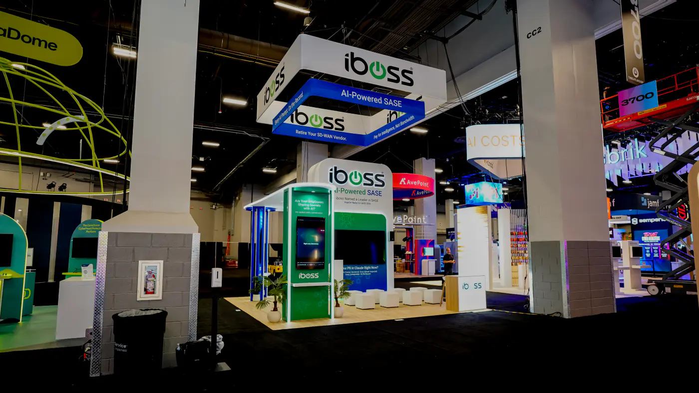 Wide view of the iboss AI-Powered SASE island booth at Black Hat USA 2026 with square hanging sign, theater screen wall, green kiosk and cube seating on light wood flooring