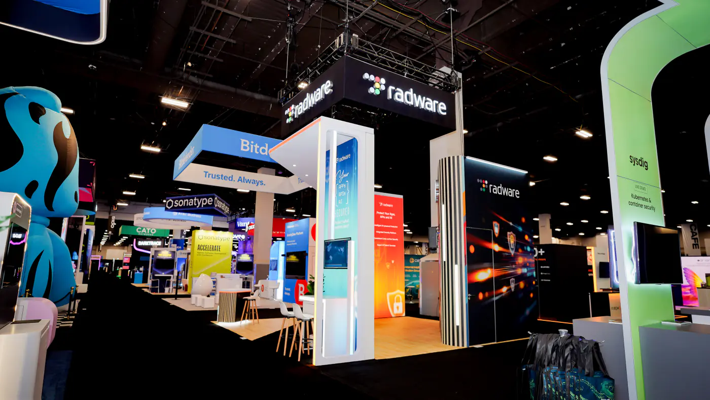 Aisle view of the Radware booth at Black Hat USA 2026 with the Your Apps APIs AI Secured pillar, slatted wood columns, orange lock graphic wall and hanging sign