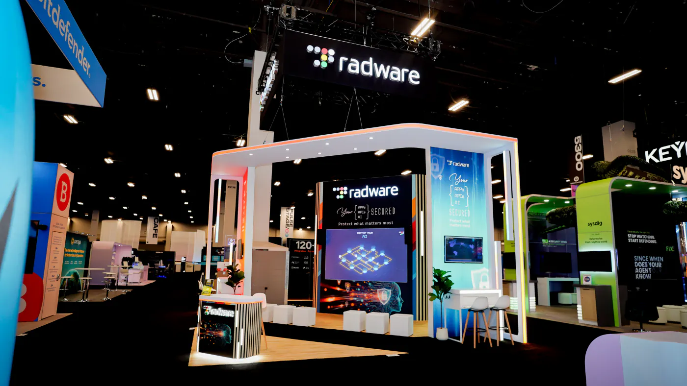Radware island booth at Black Hat USA 2026 with a black hanging sign, white portal frame, Your Apps APIs AI Secured LED wall, cube seating and a branded reception counter
