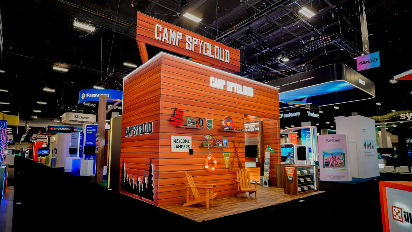 Corner view of the Camp SpyCloud cabin booth at Black Hat USA 2026 with a pine-tree silhouette end wall, camp prop shelves and Adirondack chairs