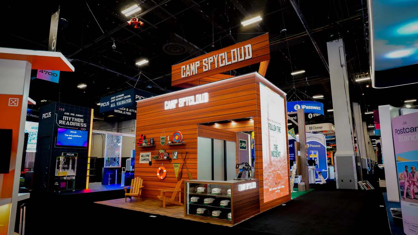 Camp SpyCloud 20x30 trade show booth at Black Hat USA 2026, a log-cabin island with lit Camp SpyCloud lettering, a Welcome Campers prop wall, Adirondack chairs and a glass hat display case