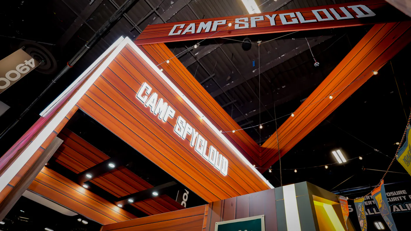 Low-angle detail of the Camp SpyCloud wood-grain hanging sign and lit cabin roofline with string lights at Black Hat USA 2026