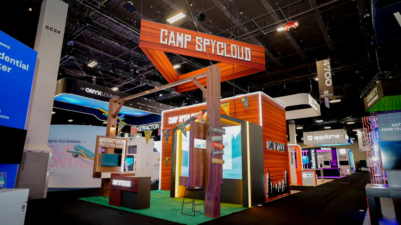 Camp SpyCloud island booth with triangular hanging sign, timber pergola and Workforce Threat Protection Cabin demo station