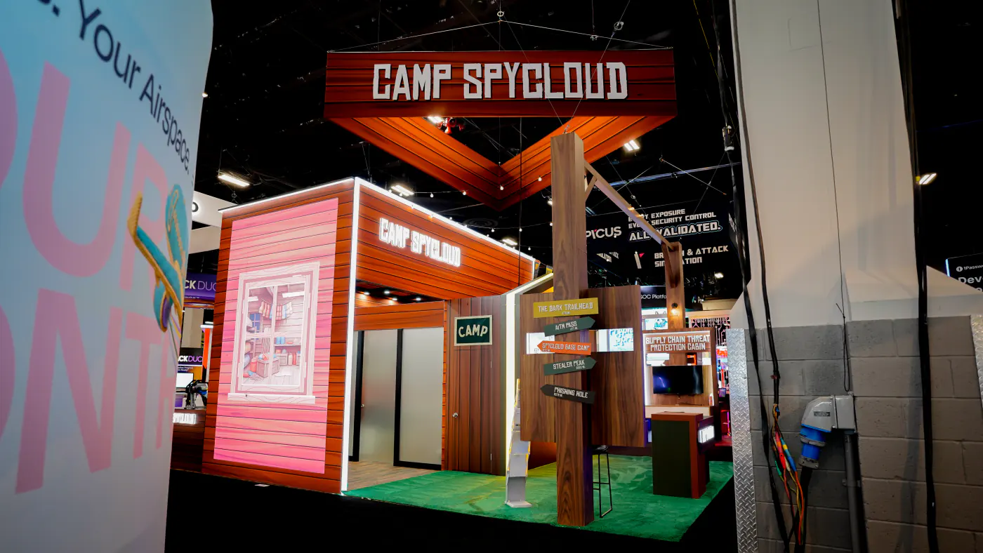Camp SpyCloud booth entrance with a wooden trail signpost pointing to Dark Trailhead, AiTM Pass, SpyCloud Base Camp, Stealer Peak and Phishing Hole